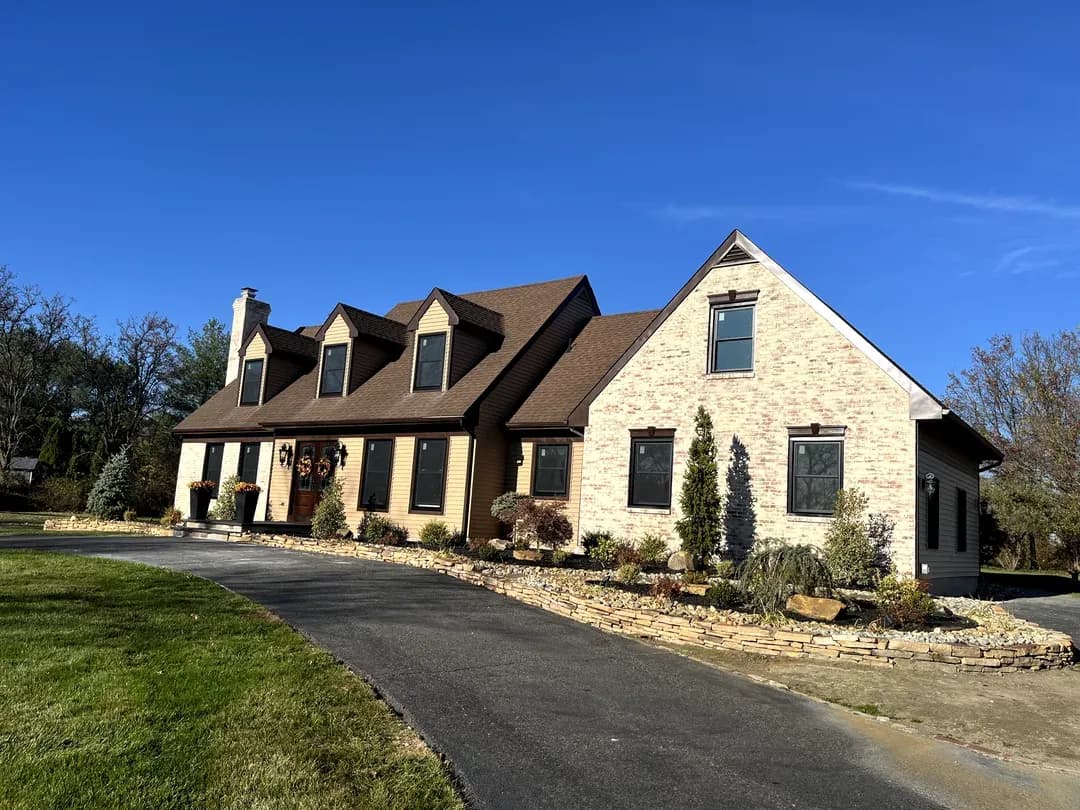 A charming suburban home with a tan brick facade, brown roof, and large dormer windows. A curved driveway and landscaped garden enhance its welcoming appearance.
