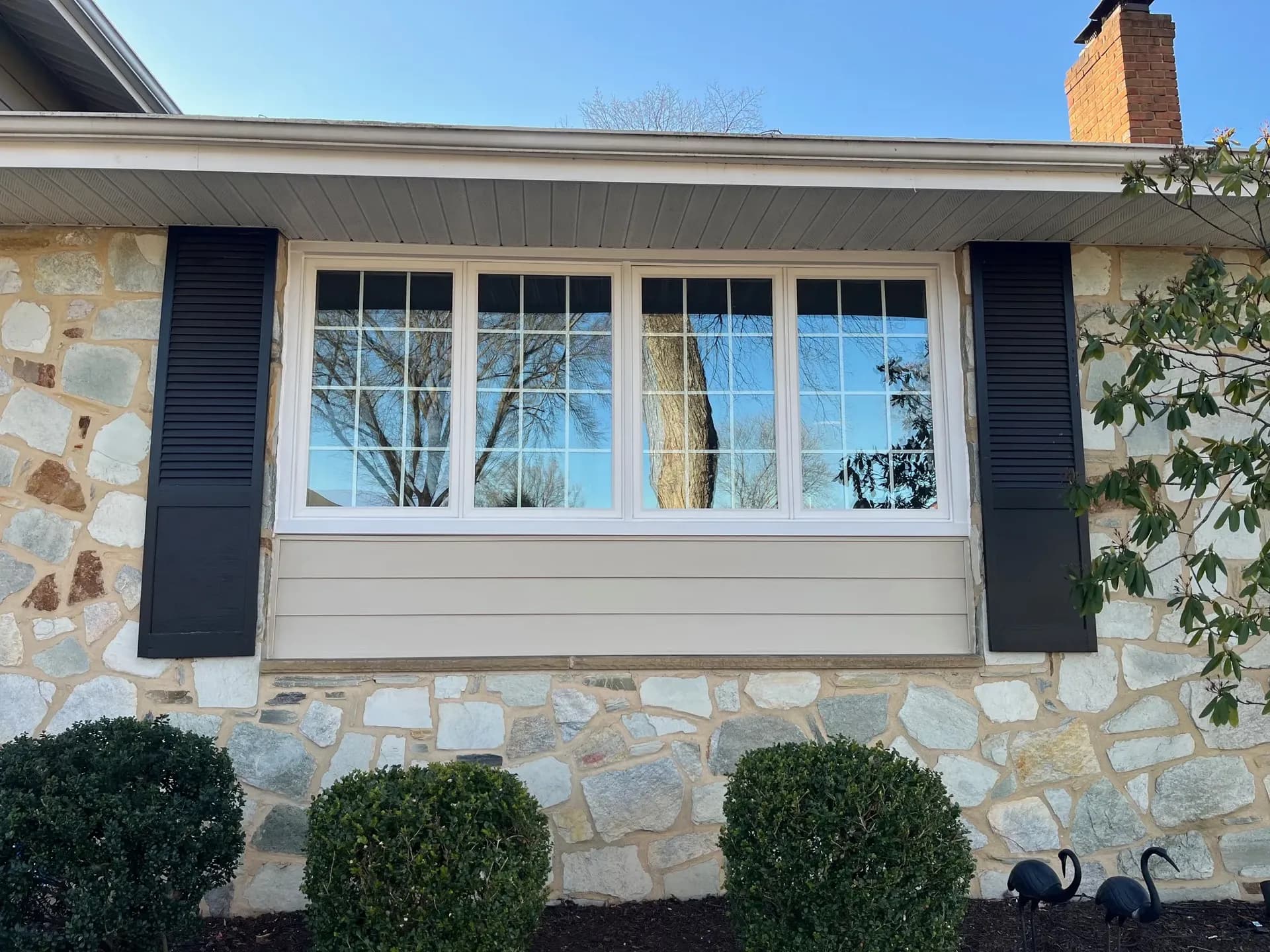 House exterior features a large window with black shutters. Reflections in the glass show trees and a blue sky. Stone facade and shrubs add charm.